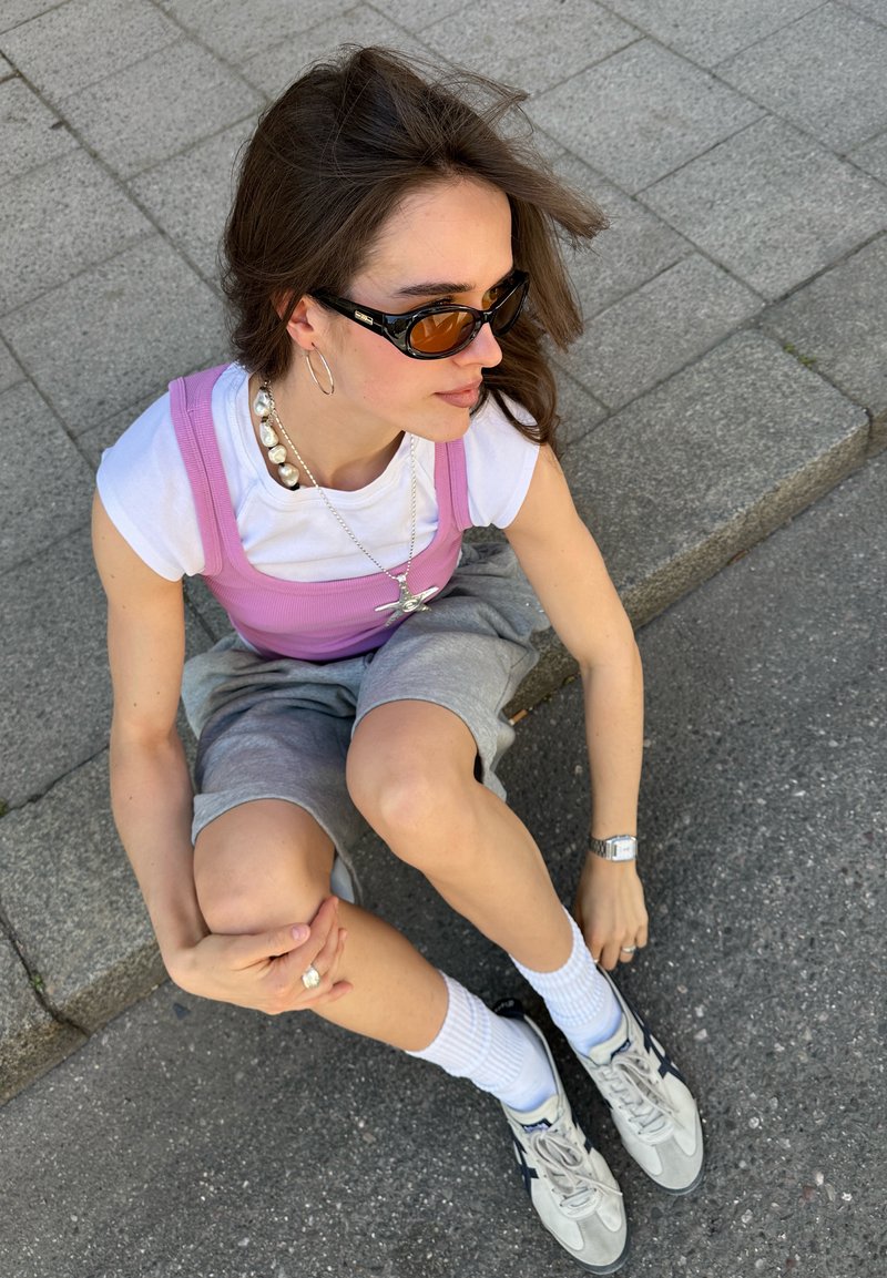 Jeune femme aux cheveux bruns portant des lunettes de soleil, une chemise blanche avec un débardeur rose, un short gris, des chaussettes blanches et des baskets, assise sur des marches en pavé.