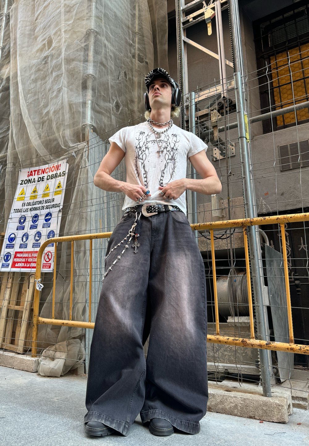 Jeans largos e pretos com uma textura desbotada, uma t-shirt gráfica, correntes em camadas e auscultadores; fundo de construção urbana.