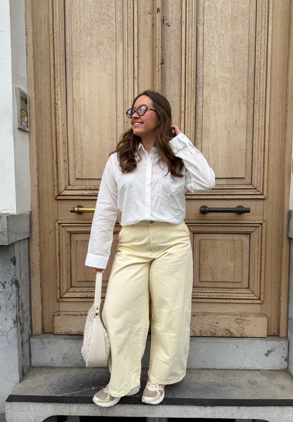 Jauna moteris su akiniais, dėvinti baltą marškinių ir kreminius kelnes, šypsodamasi stovi prie didelių medinių durų, laikydama baltą rankinę.