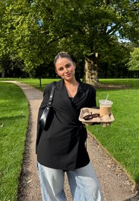 Svart omslagbluse over en svart topp, kombinert med lyseblå vide jeans. Holder en kaffekopp og solbriller på et fat. Grønn parkbakgrunn.