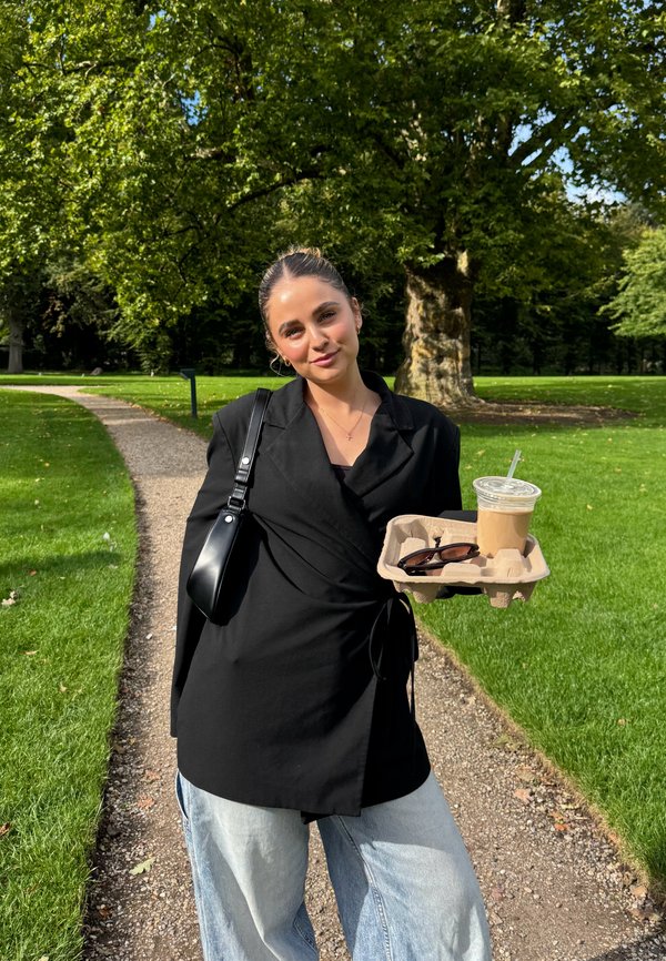 Svart omslagbluse over en svart topp, kombinert med lyseblå vide jeans. Holder en kaffekopp og solbriller på et fat. Grønn parkbakgrunn.