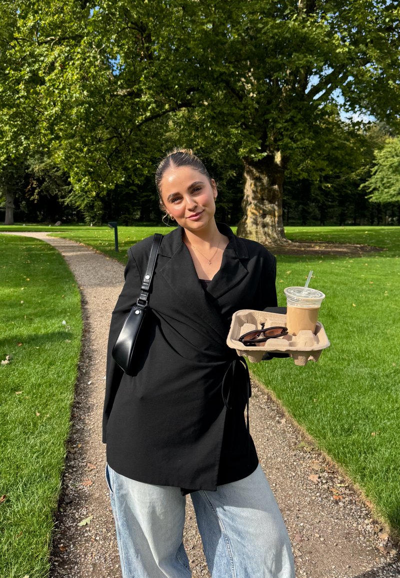 Zwarte wikkelblouse over een zwarte top, gecombineerd met lichtblauwe wijde jeans. Houdt een koffiekop en een zonnebril op een dienblad vast. Groene parken achtergrond.