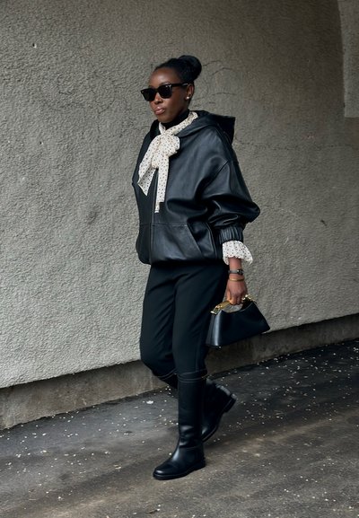 Mujer caminando con chaqueta de cuero negra, pantalones negros, botas hasta la rodilla, blusa de lunares con un lazo, gafas de sol y llevando un bolso negro.