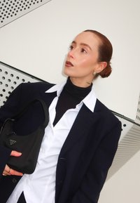 Svart Prada axelväska, strukturerad blazer, vit skjorta och svart turtleneck. Slät textur, neutrala färger och subtila smyckesdetaljer.