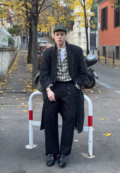 Joven con camisa de cuadros, abrigo largo oscuro, gorra plana y pantalones negros de pierna ancha sentado en una barrera metálica roja y blanca en una calle otoñal.
