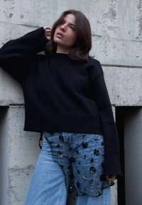 Femme aux cheveux bruns mi-longs, portant un pull noir et un jean bleu orné, debout contre un mur en béton texturé.