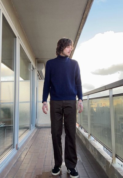 Suéter de cuello alto azul marino y pantalones rectos marrones combinados con zapatillas negras. De pie en un balcón con vista a nubes y edificios.
