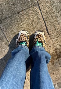 Schuhwerk mit Leopardenmuster, strukturierter Oberseite, schwarzen Akzenten und grünen Schnürsenkeln, kombiniert mit blauen Jeans, präsentiert auf einer steinernen Oberfläche.