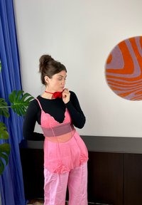 Różowy, warstwowy zestaw bielizny z przezroczystą górą i koronkowymi akcentami, zestawiony z różowymi spodniami o szerokich nogawkach. Pod spód noszona czarna bluzka z długim rękawem.