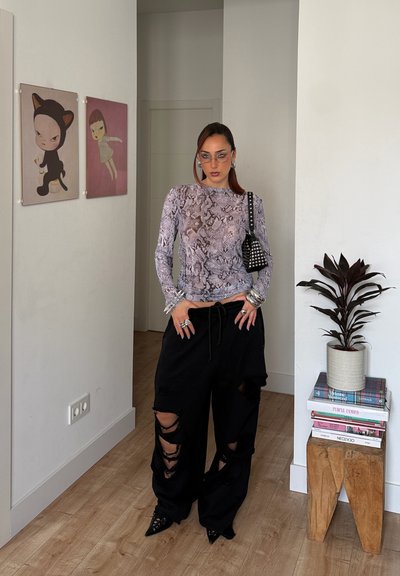 Mujer con blusa de estampado de serpiente, pantalones negros rotos, bolso con tachuelas y joyería plateada, de pie en el interior cerca de cuadros y una planta sobre libros apilados.