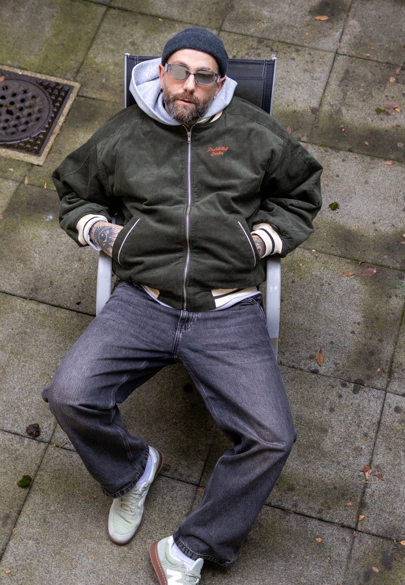 Veste bomber en velours côtelé vert avec des poignets blancs, jean gris, et baskets vert clair. La personne est assise sur une chaise en plein air.