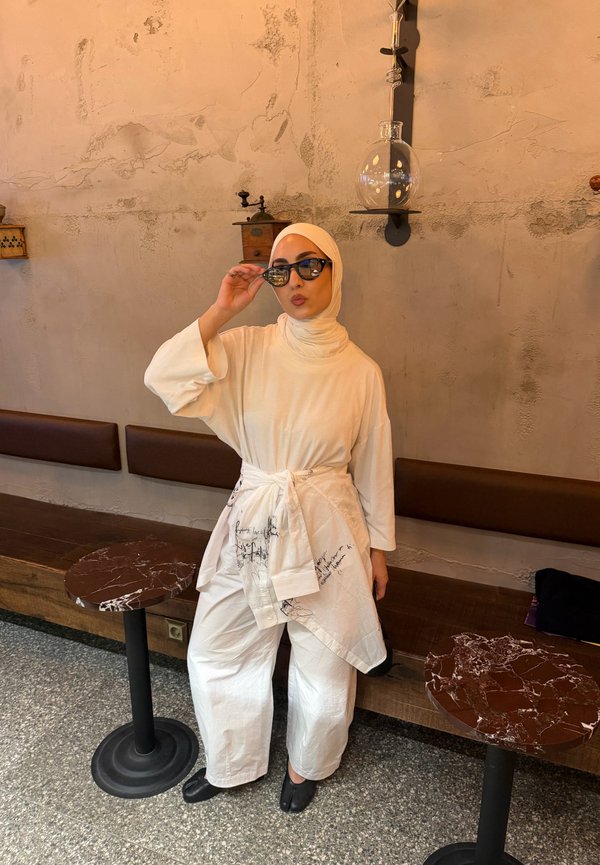 Kvinde iført hvidt tøj og hijab justerer solbriller, mens hun står ved siden af en brun bænk og to små runde borde i en café.