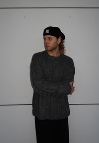 Grauer Kabelstrickpullover mit gerippten Bündchen, getragen über schwarzen Hosen, begleitet von einer schwarzen Beret mit einem weißen Logo, vor einer schlichten Wand.