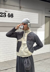 Leichte Jacke mit grauen und creamfarbenen Panels, ausgestattet mit einem hohen Kragen, einem Reißverschluss und zwei Fronttaschen. Getragen mit einer Mütze und Kopfhörern.