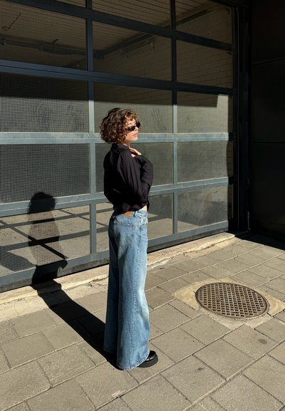 Camisa negra de manga larga, jeans sueltos de color azul claro y zapatos negros. De pie contra una pared texturizada con un patrón de rejilla, con sombras proyectadas.