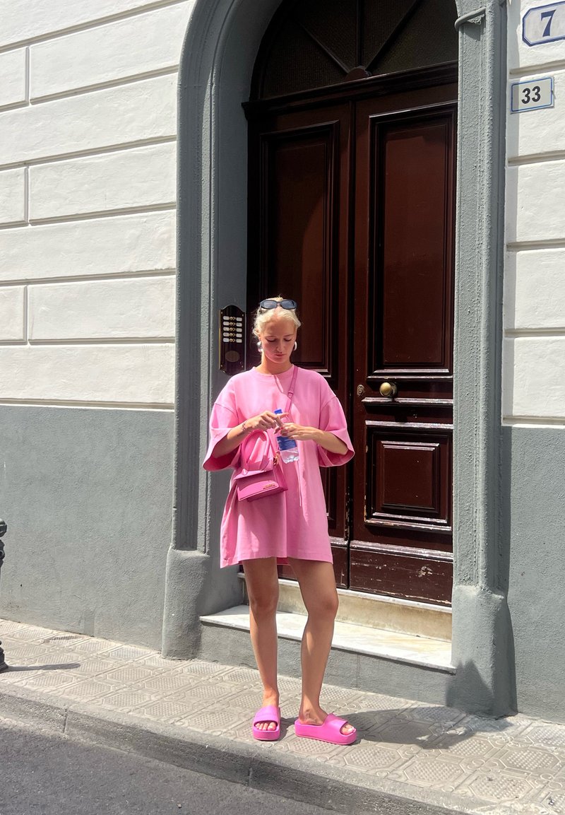Oversized ροζ φόρεμα t-shirt, ροζ τσάντα ώμου, κρατώντας μια μπουκάλα νερό, φορώντας ροζ παντόφλες, στέκεται δίπλα σε μια ξύλινη πόρτα.
