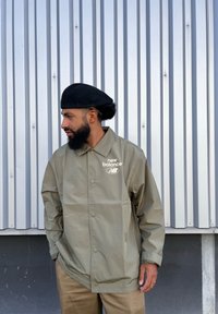 Ελαφρύ μπουφάν ελιάς με κουμπιά προσκόλλησης, κεντημένο λογότυπο "new balance" και γιακά, συνδυασμένο με καφέ παντελόνι.