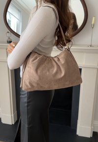 Γυναίκα που φοράει λευκό ριμπ μπλουζάκι και μαύρη φούστα, κρατώντας μια μεγάλη μπεζ τσάντα ώμου από σουέτ, σε έναν σύγχρονο, μίνιμαλ εσωτερικό χώρο.