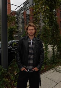 Svart skinnjakke over en grå og svart rutete skjorte, mørke bukser, med en bakgrunn av urban arkitektur og grøntområder.