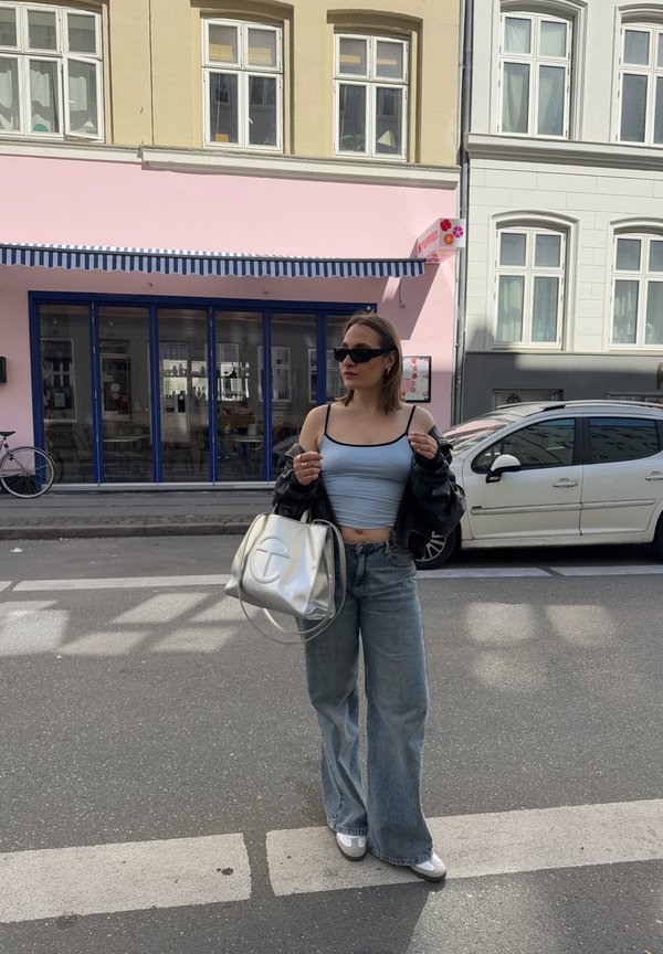 Levélkék kék crop top, fekete oversize dzseki, fakó bő szárú farmernadrág, ezüst színű táska logóval, fehér sportcipő, kültéren, városi környezetben.