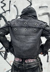 Μαύρο τζάκετ από τζιν με φθαρμένες άκρες, ασημένιες οπές και κοντή σχεδίαση, φορεμένο πάνω από μαύρο παντελόνι με ροζ φυλετικά μοτίβα.