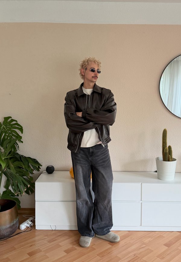 Jeune homme aux cheveux blonds bouclés et lunettes de soleil, les bras croisés, portant une veste noire et un jean ample dans une pièce minimaliste.