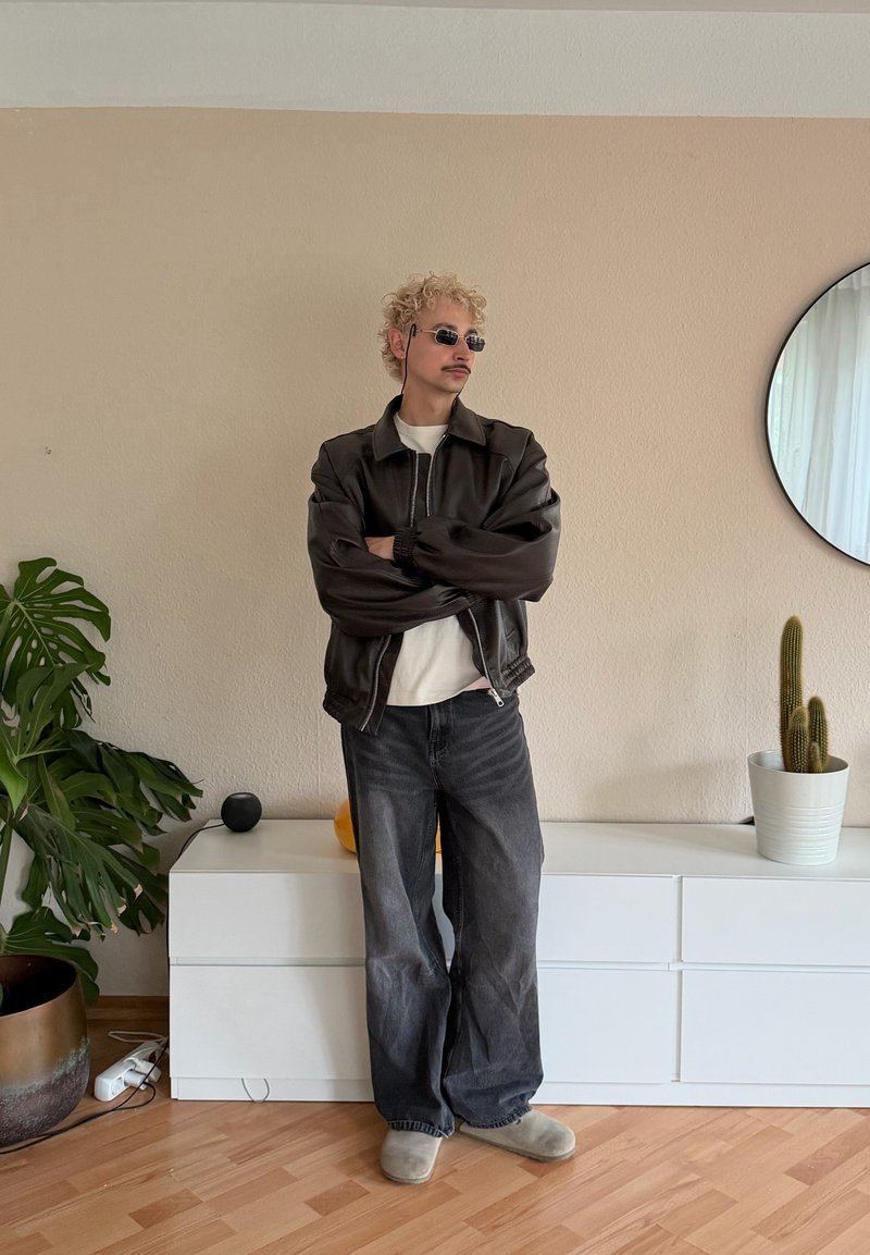 Jeune homme aux cheveux blonds bouclés et lunettes de soleil, les bras croisés, portant une veste noire et un jean ample dans une pièce minimaliste.