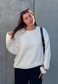 Γυναίκα με μακριά καστανά μαλλιά, που φοράει λευκό πλεκτό πουλόβερ, μαύρο παντελόνι, γυαλιά ηλίου στο κεφάλι και καφέ τσάντα ώμου, δίπλα σε γκρι τσιμεντένιο τοίχο.