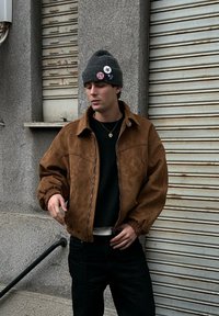 Veste en daim marron avec fermeture éclair à l’avant, large col et poignets élastiques, associée à un pull noir et un jean, sur un fond gris.