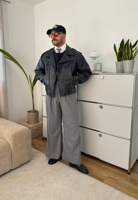 Μαύρο δερμάτινο μπουφάν μοτοσικλέτας, γκρι παντελόνι με φαρδιά γραμμή, μαύρα παπούτσια, γυαλιά, μαύρο καπέλο, στέκεται δίπλα σε ένα φυτό και μια μονάδα αποθήκευσης.