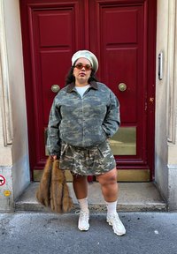 Kamakallinen takki ruskealla turkiskauluksella, samaan camo-kuosiin sopiva laskostettu hame, valkoinen t-paita, valkoiset tennarit ja kerma värinen baskeri. Ruskea turkislaukun yksityiskohta.