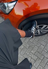 Μια μαύρη φούστα καταλήγει σε ένα πόδι που ξεκουράζεται σε μια ρόδα αυτοκινήτου. Το πόδι φοράει μαύρα και λευκά αθλητικά παπούτσια. Το αυτοκίνητο είναι πορτοκαλί με κομψά φώτα.