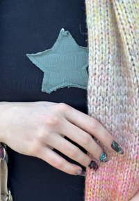 Haut noir côtelé avec patch étoile verte, et une écharpe tricotée aux teintes beige, rose et jaune ; main aux ongles multicolores reposant sur l'écharpe.