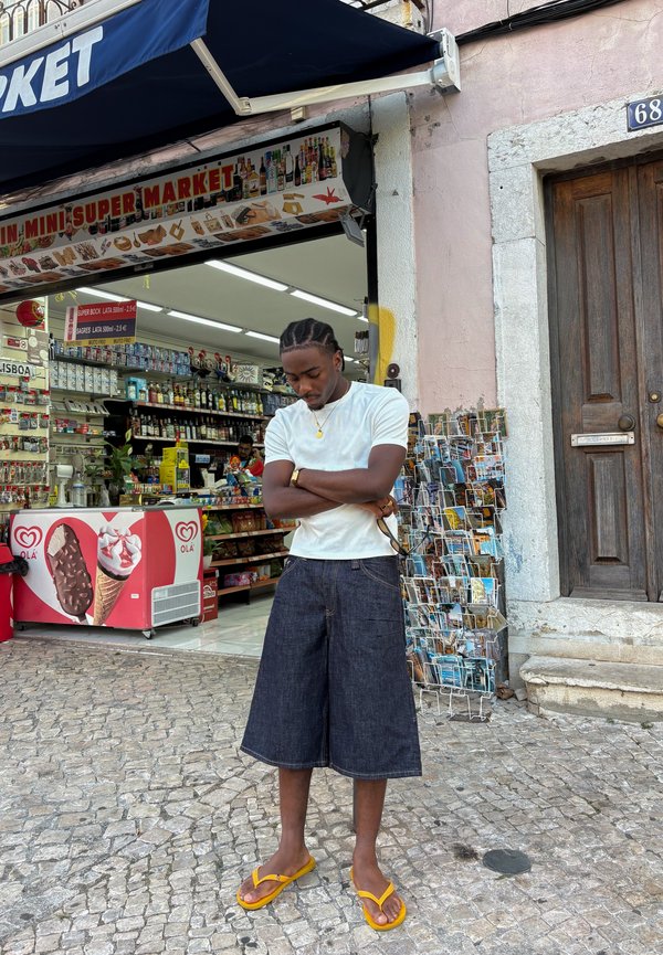 Uomo con i capelli intrecciati in piedi con le braccia incrociate fuori da un mini supermercato su una strada acciottolata, indossa una camicia bianca, pantaloncini scuri e sandali gialli.