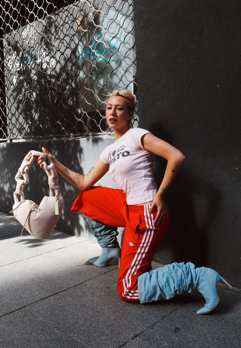 Femme blonde à genoux près d'un mur noir, portant un t-shirt court blanc, un pantalon rouge Adidas, des bottes bleues souples, tenant un sac doux beige en forme de cœur.