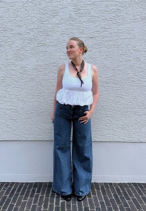 Top blanco sin mangas con dobladillo con volantes, combinado con jeans de mezclilla de pierna ancha en azul oscuro. Accesorios con una bufanda de patrón negro y marrón.