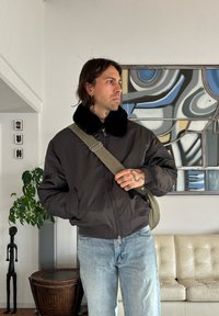 Veste bomber noire avec un col en fausse fourrure, portée sur un jean bleu clair. Un sac à bandoulière vert est porté, avec des plantes d'intérieur et des œuvres d'art en arrière-plan.