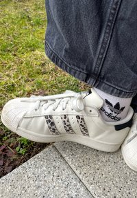 Fehér bőrsneaker három leopárdmintás csíkkal, texturált orrész és fekete díszítések. Szürke farmerrel és csíkos zoknival viselve.