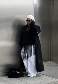Femme portant un béret blanc, des lunettes de soleil noires, un long manteau noir, un haut noir à franges et un pantalon large blanc, debout contre un mur métallique avec un sac noir à proximité.