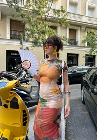 Vestido con estampado floral en tonos naranja, amarillo y verde; tejido ligero. Presenta pendientes de flores amarillas, un abanico redondo de espejo y un bolso blanco.