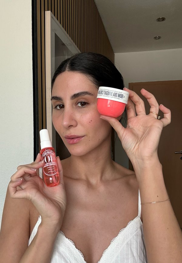 Femme aux cheveux foncés portant un haut blanc tient un pot rose étiqueté « BOM DIA BRIGHT CREAM » et un petit flacon spray avec un liquide orange.