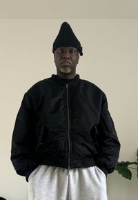 Fekete bomberdzseki elöl cipzáros, bordázott mandzsettákkal és oldalzsebekkel. Világosszürke melegítőnadrággal párosítva. Fekete kötött sapka, rövid és texturált.