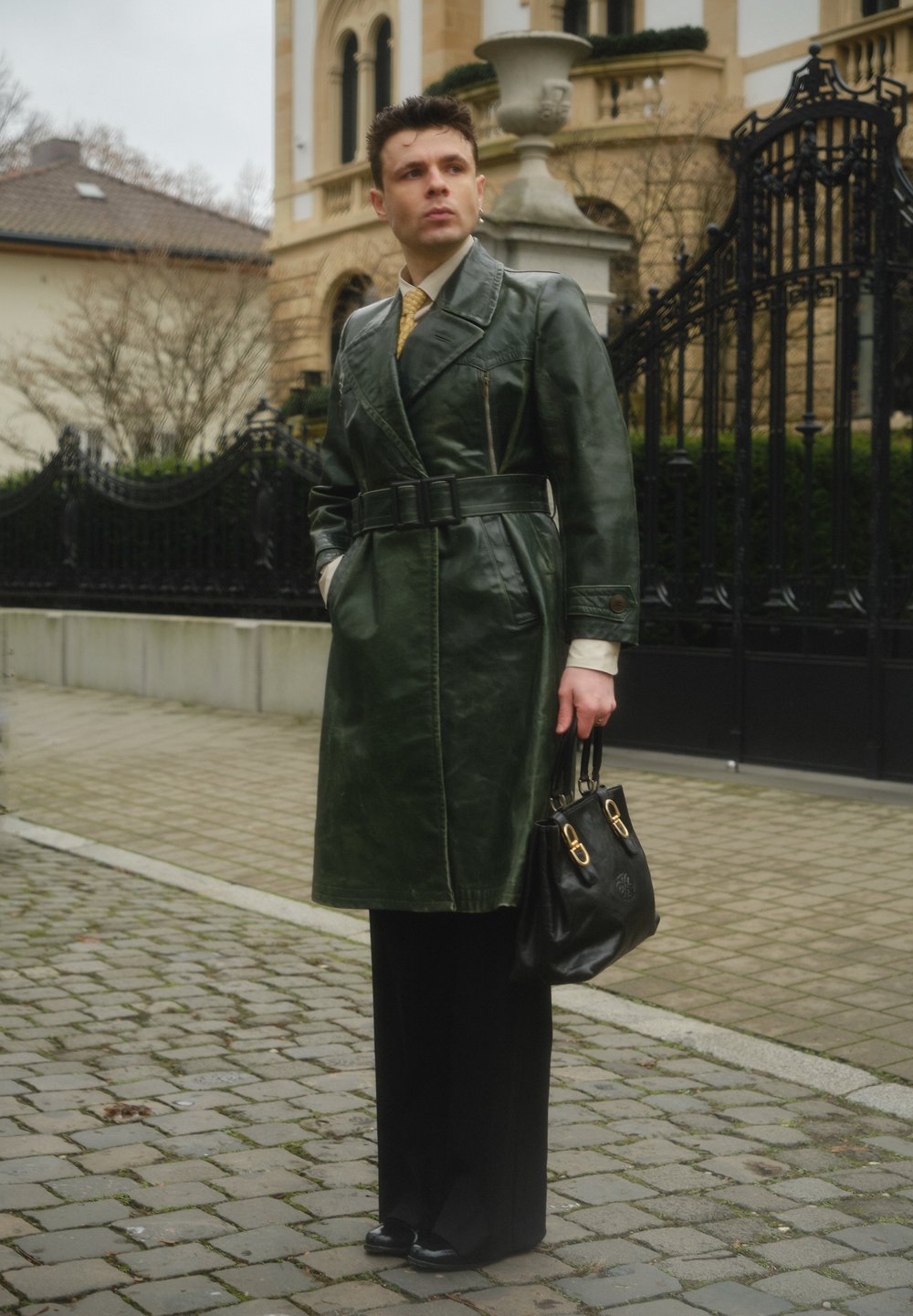 Manteau trench-coat en cuir vert avec une taille ceinturée, mettant en valeur des coutures marquées et de grandes poches. Associé à un pantalon noir et un sac à main noir.