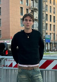 Sort strikket sweater, lyserød skjorte med krave under, falmede blå jeans og en guld hængeørering. Urban baggrund med trafikskilt.