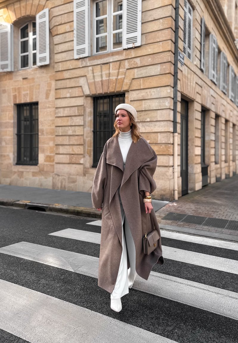 Femme portant un long manteau marron, un béret blanc et des bottes, traversant un passage piéton en ville près de bâtiments en pierre beige.