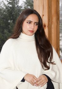 Poncho en maille blanche avec un col montant, présentant un design ample et fluide ainsi qu'un tissu texturé. Cheveux longs et foncés partiellement visibles.