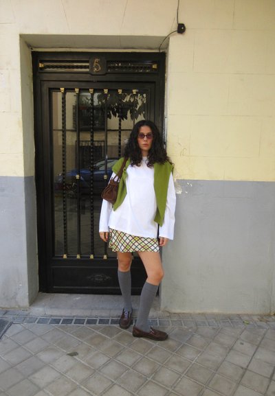 Mujer con cabello rizado que lleva gafas de sol, camisa blanca de manga larga, suéter verde sobre los hombros, falda de cuadros, calcetas grises hasta la rodilla y zapatos marrones se encuentra junto a una puerta negra.