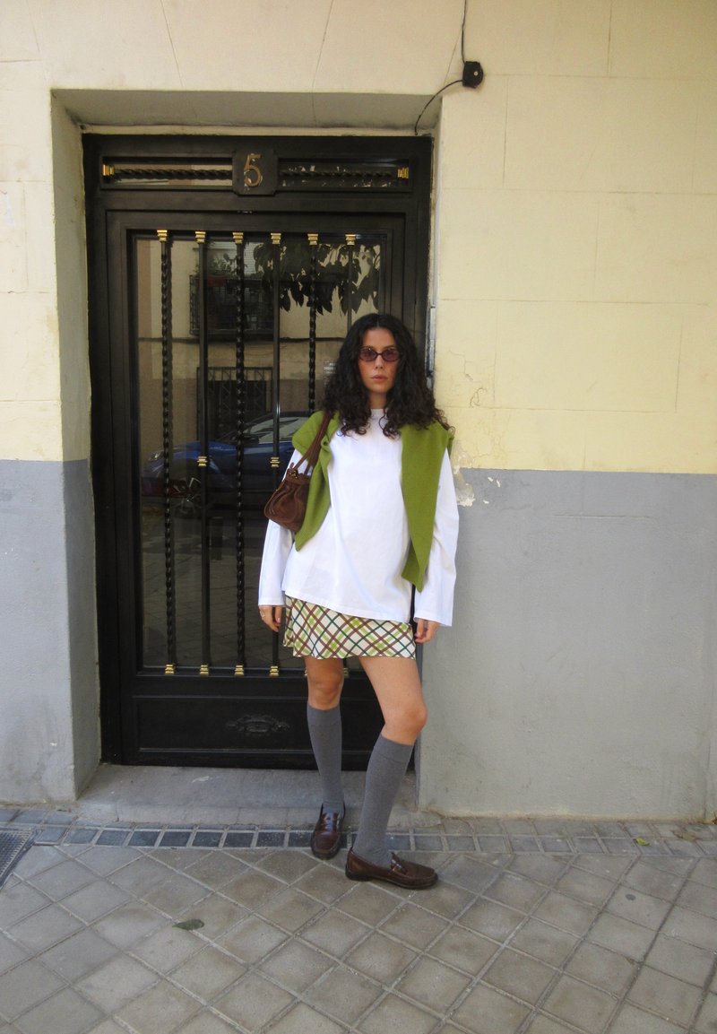 Donna dai capelli ricci che indossa occhiali da sole, una camicia bianca a maniche lunghe, un maglione verde sulle spalle, una gonna a quadri, calze grigie fino al ginocchio e scarpe marroni, sta in piedi accanto a una porta nera.