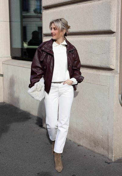 Mujer caminando por la calle de la ciudad con una chaqueta de cuero marrón oscuro, camisa blanca, pantalones blancos, botas marrones y sosteniendo un bolso blanco esponjoso.