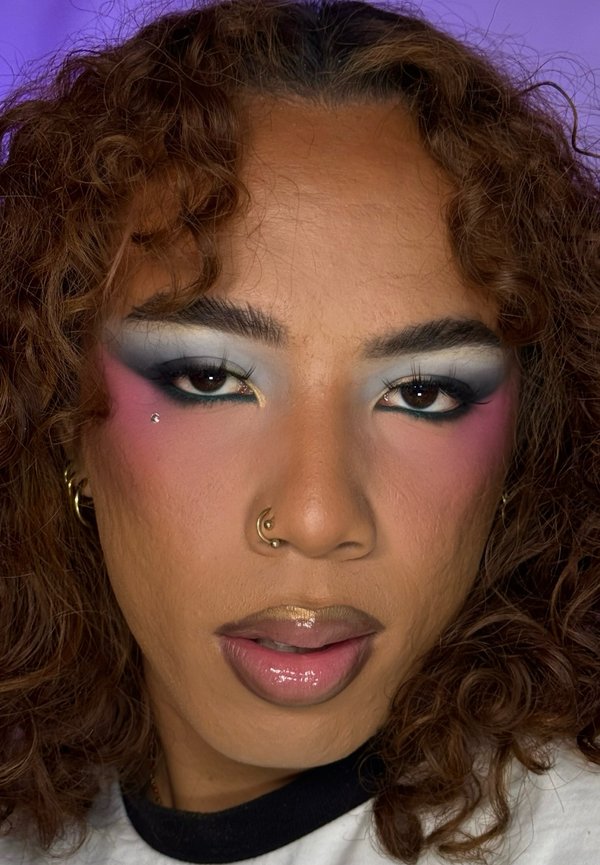 Person mit lockigem Haar, die auffälligen blauen und pinken Lidschatten, goldenen und pinken glänzenden Lippenstift, Nasenpiercing und kleinen Gesichtsschmuck nahe dem Auge trägt.